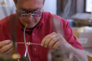 man blowing in tube handling torch and glass