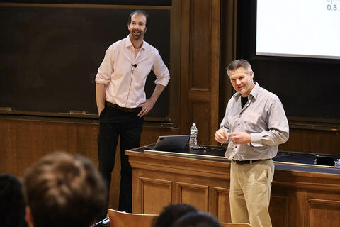 Two men speaking to and smiling at an audience