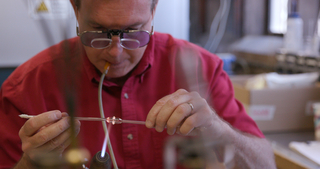 man blowing in tube handling torch and glass