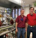 Two men standing next to a lathe in a glass shop